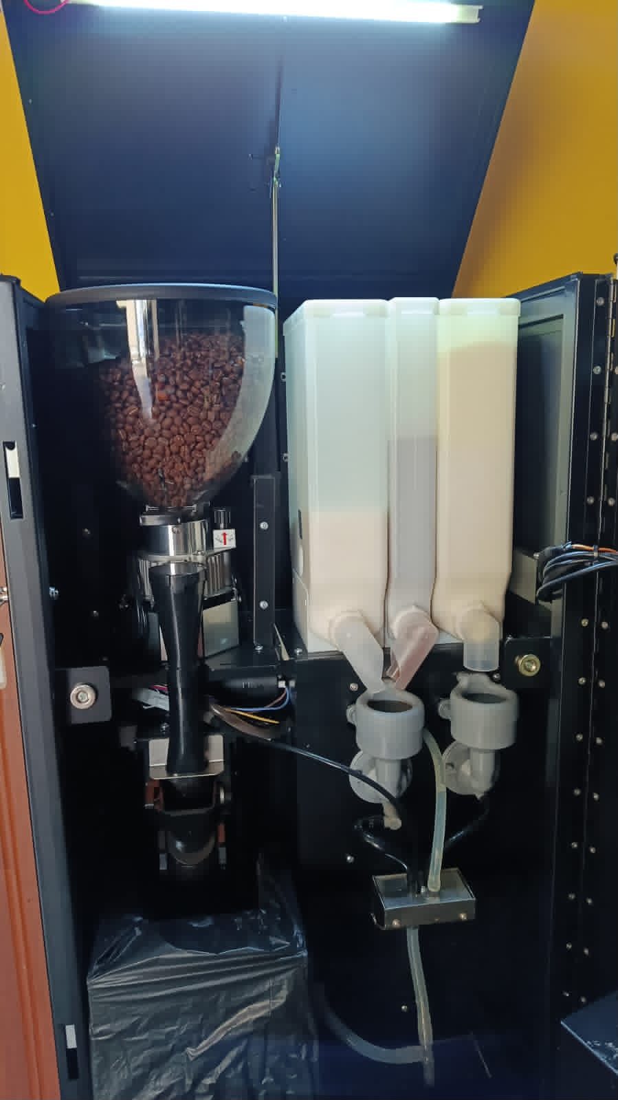 The inside of a Vendlite vending machine, showing a hopper filled with whole roasted coffee beans above the grinder, next to canisters of milk and chocolate powder